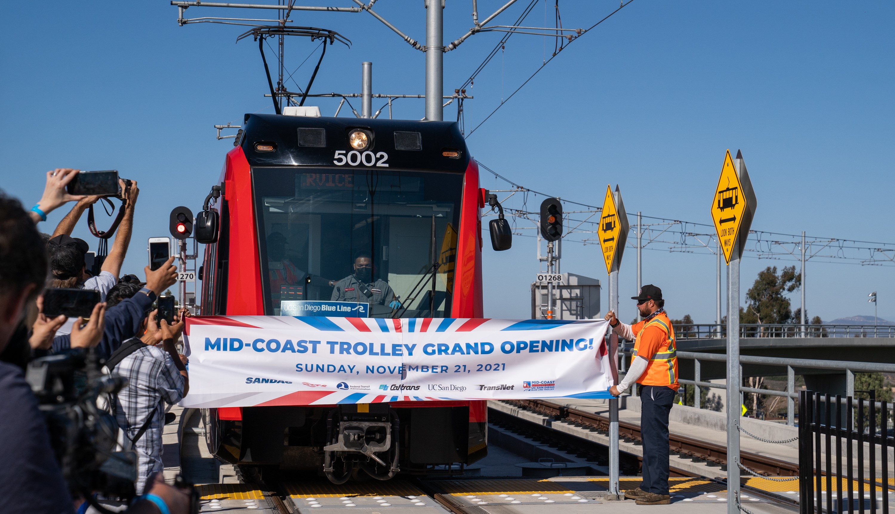 Red Trolley breaking through Mid-Coast Trolley Grand Opening banner while bystanders cheer and take pictures